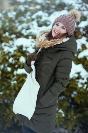 Foto de Sonriente mujer moderna de mediana edad en abrigo verde y sombrero marrón al aire libre en el parque de la ciudad en invierno con gorro sombrero cerca de ramas nevadas. - Imagen libre de derechos