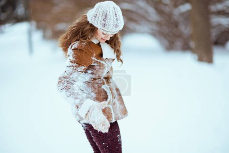 Photo for Elegant middle aged woman in brown hat and scarf in sheepskin coat with mittens shaking snow off clothes outside in the city in winter. - Royalty Free Image