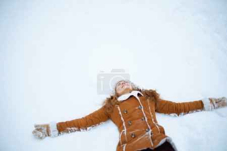 Photo for Happy stylish 40 years old woman in brown hat and scarf in sheepskin coat with mittens laying on snow outside in the city in winter. - Royalty Free Image