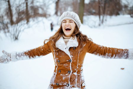Foto de Mujer elegante sonriente en sombrero marrón y bufanda en abrigo de piel de oveja con manoplas y brazos abiertos regocijándose fuera en la ciudad en invierno. - Imagen libre de derechos