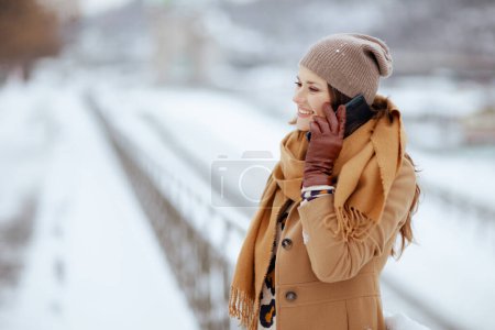 Photo for Happy elegant 40 years old woman in brown hat and scarf in camel coat with gloves talking on a smartphone outside in the city in winter. - Royalty Free Image