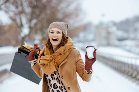 Photo for Smiling modern woman in brown hat and scarf in camel coat with gloves, shopping bags and cup of soy latte outside in the city in winter. - Royalty Free Image