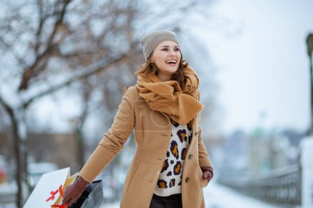 Téléchargez les photos : Heureuse femme élégante en chapeau marron et écharpe en manteau de chameau avec gants et sacs à provisions à l'extérieur de la ville en hiver. - en image libre de droit