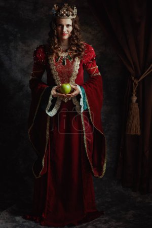 Photo for Full length portrait of medieval queen in red dress with green apple and crown on dark gray background. - Royalty Free Image