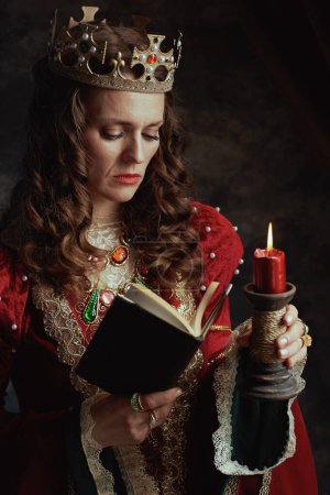 Foto de Reina medieval en vestido rojo con libro, vela y corona sobre fondo gris oscuro. - Imagen libre de derechos