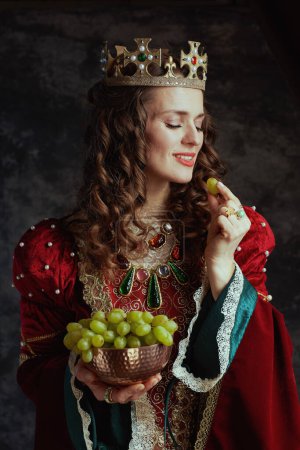 Téléchargez les photos : Reine médiévale souriante en robe rouge avec assiette de raisins et couronne sur fond gris foncé. - en image libre de droit
