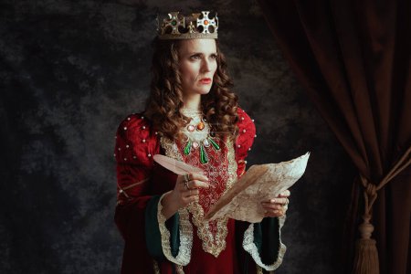Téléchargez les photos : Reine médiévale coûteuse en robe rouge avec parchemin et couronne sur fond gris foncé. - en image libre de droit