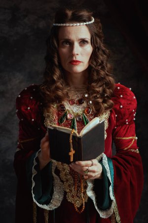 Foto de Pensativa reina medieval en vestido rojo con libro y rosario sobre fondo gris oscuro. - Imagen libre de derechos