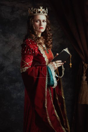 Foto de Reina medieval en vestido rojo con libro, rosario y corona sobre fondo gris oscuro. - Imagen libre de derechos