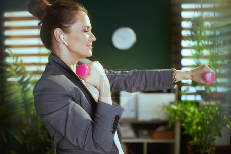 Foto de Lugar de trabajo sostenible. mujer de negocios moderna feliz en la oficina verde moderna con mancuernas y auriculares inalámbricos. - Imagen libre de derechos