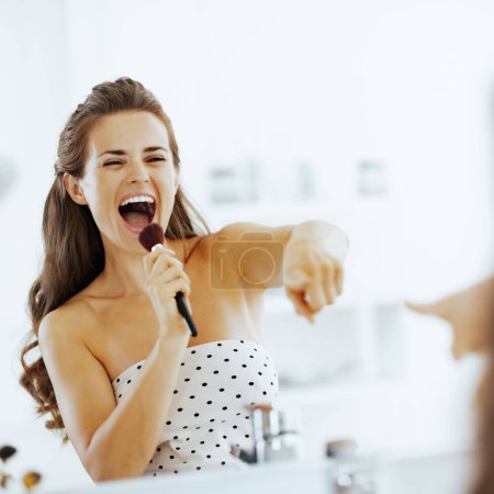Téléchargez les photos : Joyeux jeune femme chantant dans la brosse de maquillage dans la salle de bain - en image libre de droit