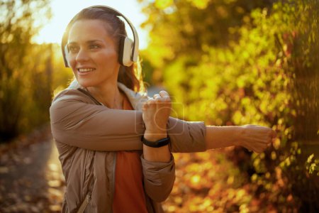 Téléchargez les photos : Bonjour l'automne. heureuse femme à la mode en vêtements de fitness dans le parc à l'écoute de la musique avec écouteurs et étirement. - en image libre de droit