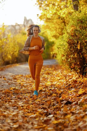 Photo for Hello autumn. Full length portrait of fit woman in fitness clothes in the park listening to the music with headphones and jogging. - Royalty Free Image