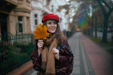 Téléchargez les photos : Bonjour l'automne. heureuse femme d'âge moyen moderne en chapeau rouge avec des feuilles d'automne et foulard marche dans la ville. - en image libre de droit