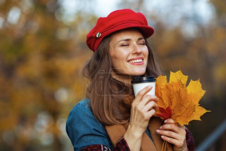 Foto de Hola otoño. mujer elegante y relajada en camisa vaquera y sombrero rojo con hojas de otoño, bufanda y café en el parque de la ciudad. - Imagen libre de derechos