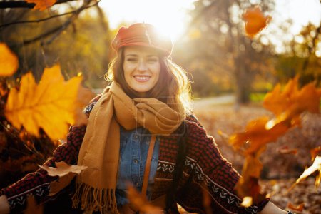 Téléchargez les photos : Bonjour l'automne. Portrait de heureuse élégante femme en chapeau rouge avec écharpe jetant des feuilles d'automne dans le parc de la ville. - en image libre de droit