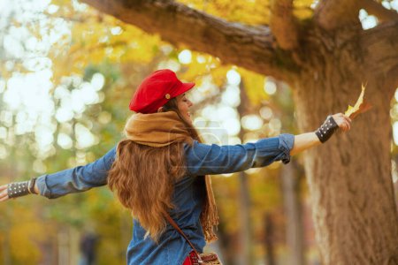 Foto de Hola otoño. mujer de moda feliz en jeans camisa y sombrero rojo con hoja de otoño, bufanda y guantes regocijándose en el parque de la ciudad. - Imagen libre de derechos