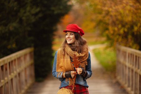 Photo for Hello autumn. smiling elegant woman in jeans shirt and red hat with autumn leaf, scarf and gloves in the city park. - Royalty Free Image
