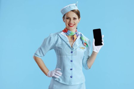 Foto de Sonriente elegante azafata femenina aislada sobre fondo azul en uniforme azul mostrando la pantalla en blanco del smartphone. - Imagen libre de derechos