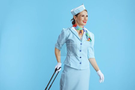 Foto de Mujer auxiliar de vuelo elegante sonriente contra fondo azul en uniforme azul con bolsa de carro mirando a la distancia. - Imagen libre de derechos