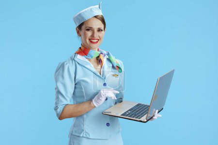 Téléchargez les photos : Souriant élégante hôtesse de l'air féminine sur fond bleu en uniforme bleu en utilisant un ordinateur portable. - en image libre de droit