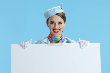 Téléchargez les photos : Hôtesse de l'air moderne souriante sur fond bleu uniforme bleu montrant tableau blanc. - en image libre de droit