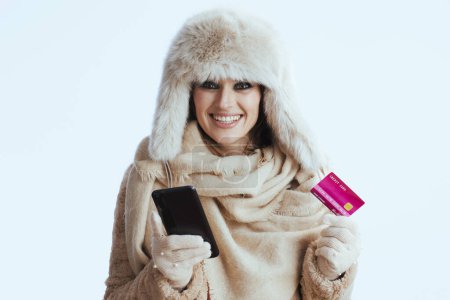 Foto de Feliz mujer moderna de mediana edad en abrigo de invierno y sombrero de piel sobre fondo blanco en guantes blancos con tarjeta de crédito y teléfono inteligente. - Imagen libre de derechos