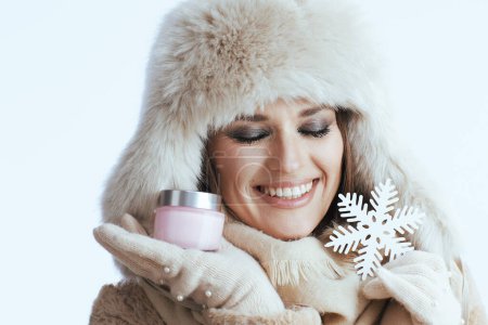 Photo for Smiling stylish 40 year old woman in winter coat and fur hat against white background in white gloves with facial cream and snowflake. - Royalty Free Image