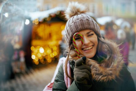 fröhliches modernes Weibchen in grünem Mantel und braunem Hut auf dem Wintermarkt in der Stadt mit Zuckerrohr.