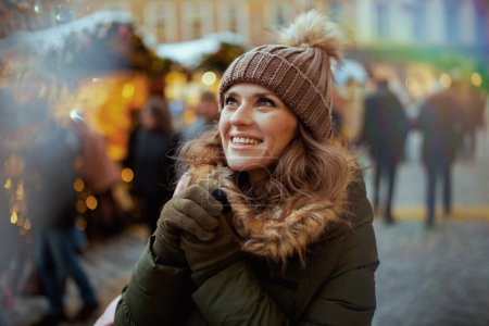 Foto de Sonriente joven hembra en abrigo verde y sombrero marrón en la feria de Navidad en las manos de calentamiento de la ciudad. - Imagen libre de derechos
