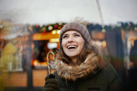 Foto de Mujer moderna feliz en abrigo verde y sombrero marrón en la feria de Navidad en la ciudad con bastón de caramelo. - Imagen libre de derechos