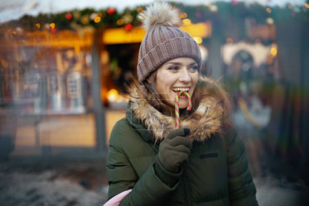 Foto de Feliz 40 años de edad, mujer de abrigo verde y sombrero marrón en la feria de invierno en la ciudad con bastón de caramelo. - Imagen libre de derechos