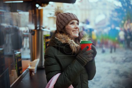 Foto de Mujer joven feliz en abrigo verde y sombrero marrón en la feria de Navidad en la ciudad con bebida de Navidad. - Imagen libre de derechos