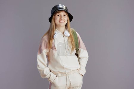 Photo for Portrait of smiling trendy teen girl in beige tracksuit with backpack, headphones and hat isolated on grey background. - Royalty Free Image
