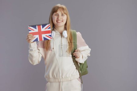 Téléchargez les photos : Portrait de fille tendance souriante en survêtement beige avec sac à dos, classeur drapeau anglais et écouteurs isolés sur fond gris. - en image libre de droit