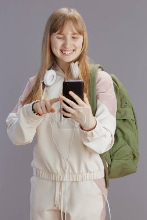 Photo for Happy trendy pupil in beige tracksuit with backpack, headphones and smartphone against grey. - Royalty Free Image