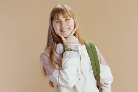 Photo for Portrait of smiling modern school girl in beige tracksuit with backpack and headphones isolated on beige background. - Royalty Free Image