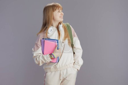 Photo for Smiling modern girl in beige tracksuit with workbooks and headphones against grey. - Royalty Free Image