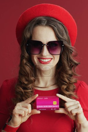 Foto de Feliz San Valentín. feliz elegante mujer de 40 años en vestido rojo y boina aislada sobre fondo rojo con tarjeta de crédito. - Imagen libre de derechos