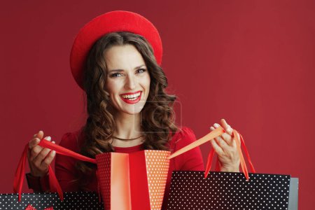 Foto de Feliz San Valentín. mujer moderna feliz en vestido rojo y boina aislada sobre fondo rojo con bolsas de compras. - Imagen libre de derechos