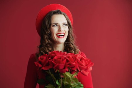 Téléchargez les photos : Joyeux Valentin. Femme élégante et souriante en robe rouge et béret sur fond rouge avec des roses rouges. - en image libre de droit