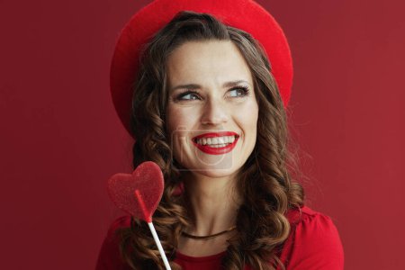 Foto de Feliz San Valentín. sonriente mujer moderna de mediana edad en vestido rojo y boina contra fondo rojo con caramelo en forma de corazón en palo. - Imagen libre de derechos