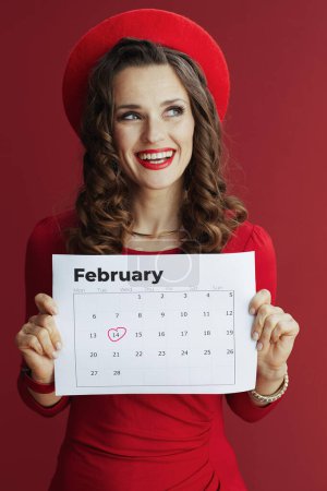 Foto de Feliz San Valentín. sonriente mujer elegante en vestido rojo y boina con calendario de febrero aislado en rojo. - Imagen libre de derechos