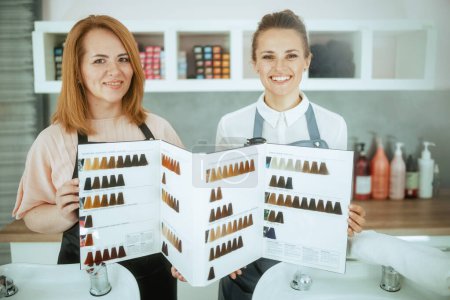 Photo for Smiling two women hairdressers in uniform in modern hair studio with hair color chart. - Royalty Free Image