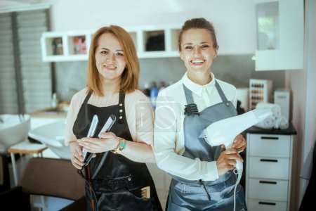 Téléchargez les photos : Portrait de deux femmes heureuses de 40 ans coiffeuses dans des tabliers dans un salon de beauté moderne avec lisseur et sèche-cheveux. - en image libre de droit