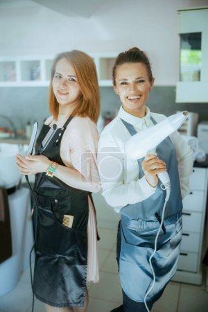Téléchargez les photos : Heureux deux femmes coiffeurs dans des tabliers dans un studio de coiffure moderne avec lisseur et sèche-cheveux. - en image libre de droit
