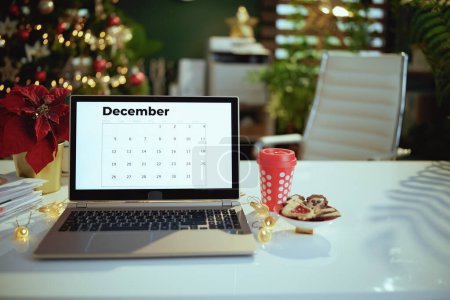 Téléchargez les photos : Bureau vert décoré de Noël moderne avec calendrier de décembre sur écran d'ordinateur portable, poinsettia, tasse rouge de boisson chaude de Noël et biscuits. - en image libre de droit