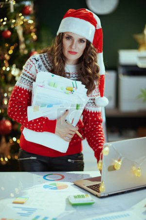 Photo for Christmas time. elegant middle aged small business owner woman in santa hat and red Christmas sweater with documents and laptop working in modern green office with Christmas tree. - Royalty Free Image