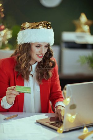 Photo for Christmas time. happy modern small business owner woman in santa hat and red jacket with laptop and credit card in modern green office with Christmas tree. - Royalty Free Image