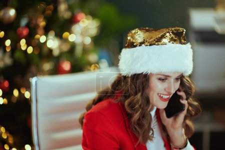 Téléchargez les photos : Noël. souriant élégant propriétaire de petite entreprise femme en santa chapeau dans le bureau vert moderne avec arbre de Noël en veste rouge parler sur un smartphone. - en image libre de droit